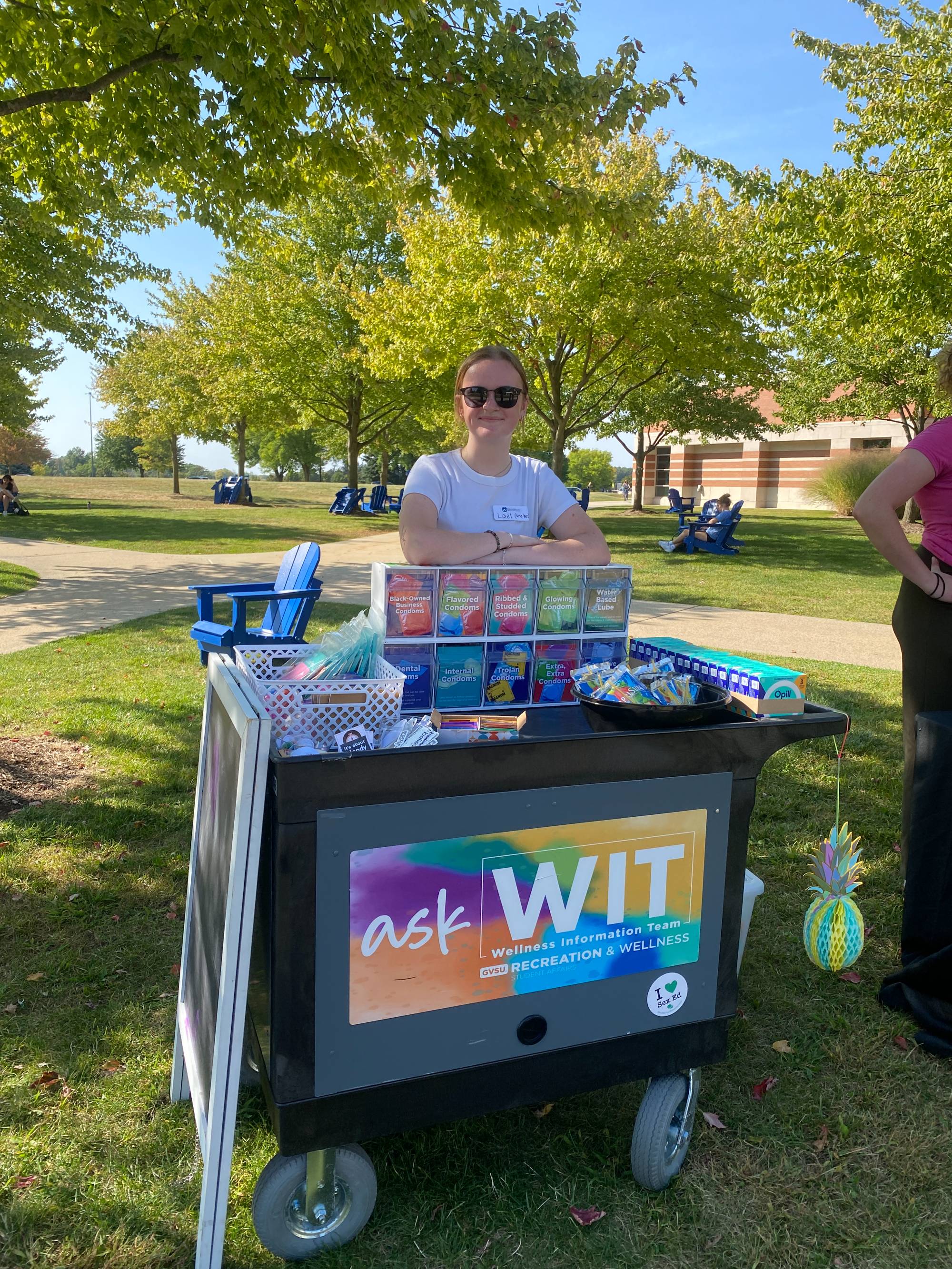 Young woman with sunglasses on standing outside behind a cart that says "Ask the Wellness Information Team"
